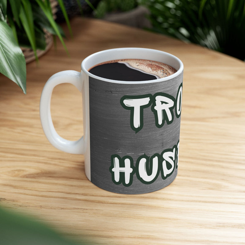 'Trophy Husband' Mug side view on a wooden table with coffee.
