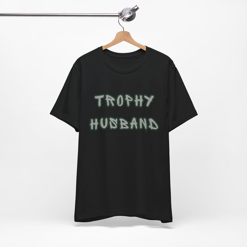 Black t-shirt with 'Trophy Husband' text on a hanger against a white background