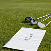Customizable white golf towel with a "your image or design" placeholder laying on the grass next to come golf clubs.