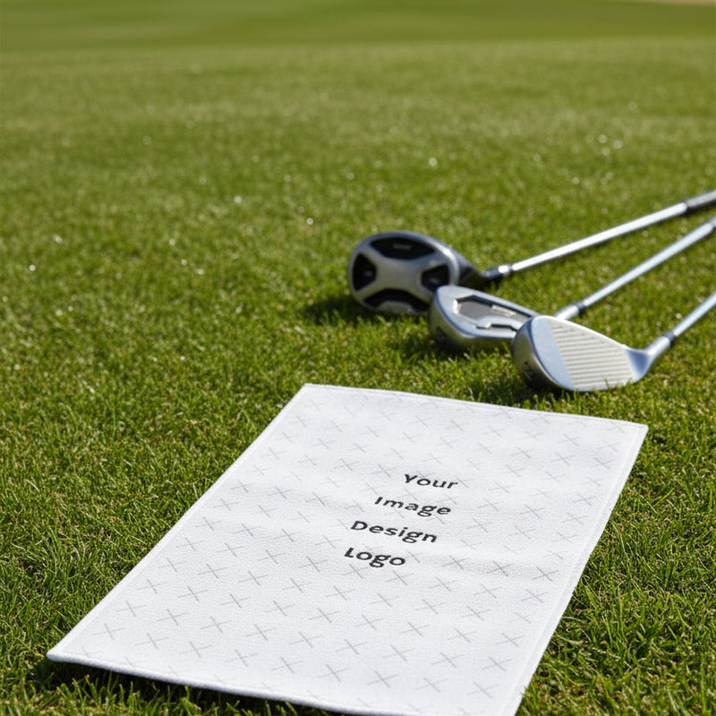 Customizable white golf towel with a "your image or design" placeholder laying on the grass next to come golf clubs.