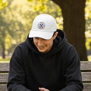 Guy wearing all white classic ball cap with a circular emblem featuring an eagle and text , along with the American flag on the side sitting on a bench.