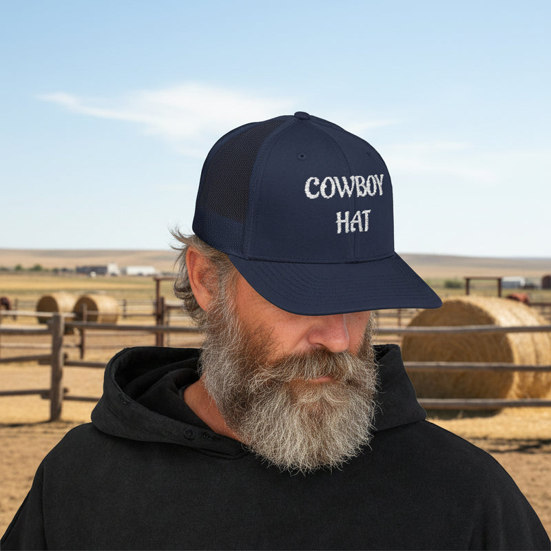 Man wearing a navy blue  trucker hat with 'Cowboy Hat' text in front of a ranch. 