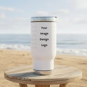 Customizable white tumbler with silver lid with a "your image or design" placeholder on a small table at the beach. 