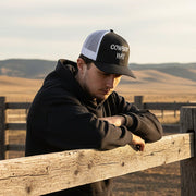 man wearing a black and white trucker hat with 'Cowboy Hat' text leaning against a fence. 