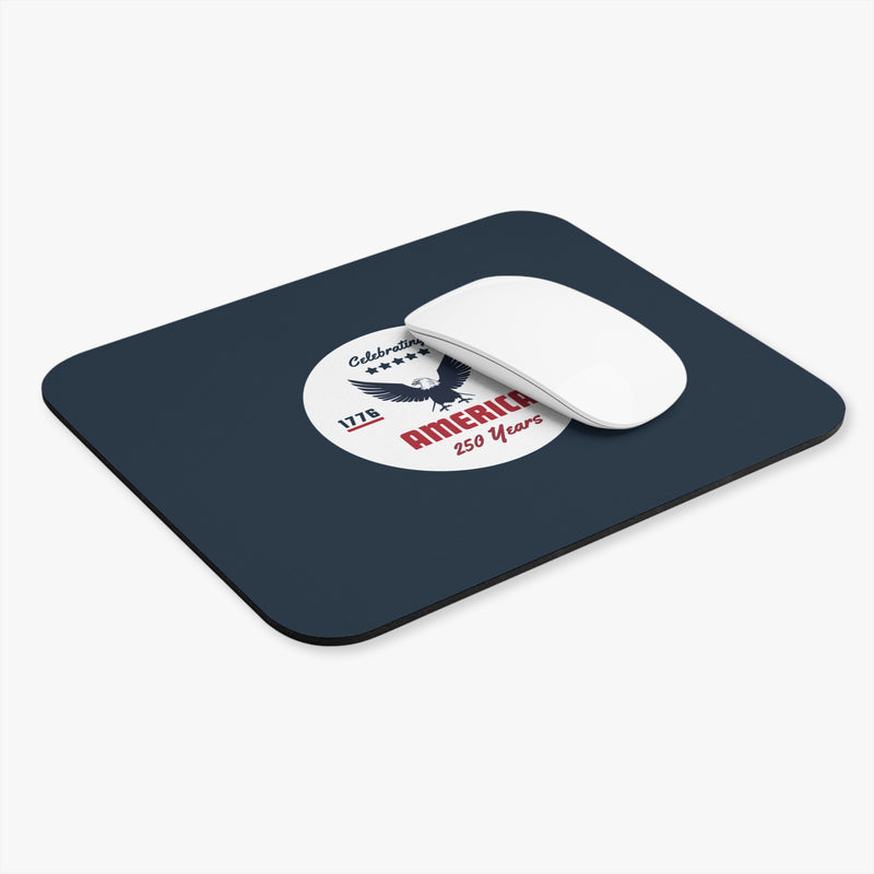Navy blue mouse pad with a circular design featuring an eagle and text celebrating America's 250th anniversary with a white computer mouse.