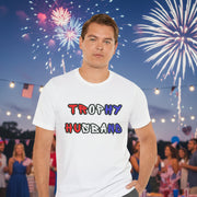 Man wearing a white t-shirt with 'Trophy Husband' in red white and blue text in front of fireworks. 