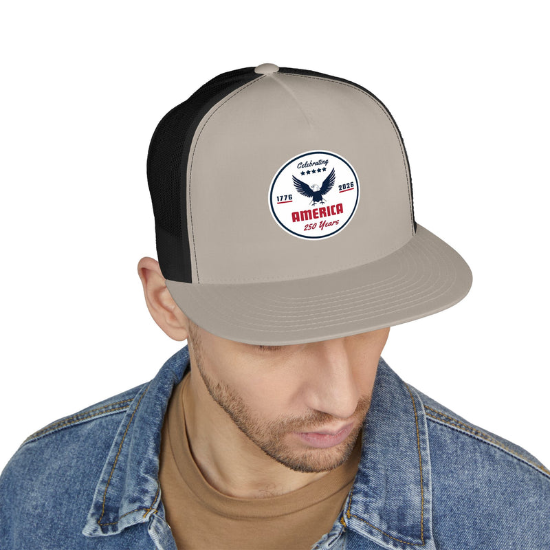 Man wearing a stone color front and black back flat bill trucker cap with a logo 'celebrating America 250 years' on a white background.
