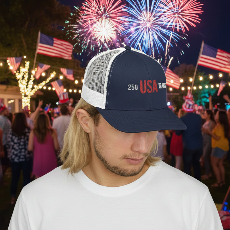 Man wearing a black and navy trucker cap with 'USA 250 Years' in front of a party. 
